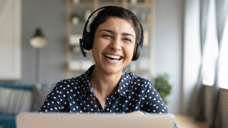 Woman wearing headphones and polka dot shirt laughing during a video call, sitting at home with laptop