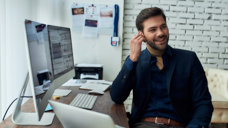 Smiling man in navy blazer and turtleneck at wooden desk with computer, against white brick wall in office
