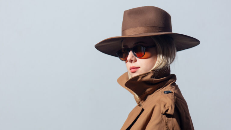 Woman wearing wide-brimmed brown hat, sunglasses, and tan trench coat against light background, profile view