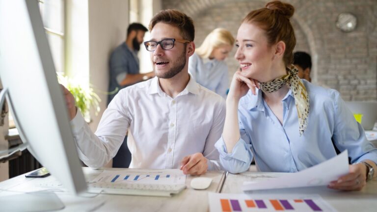 Young professionals looking at computer screen together, discussing data charts, woman with red hair in blue shirt and leopard scarf.