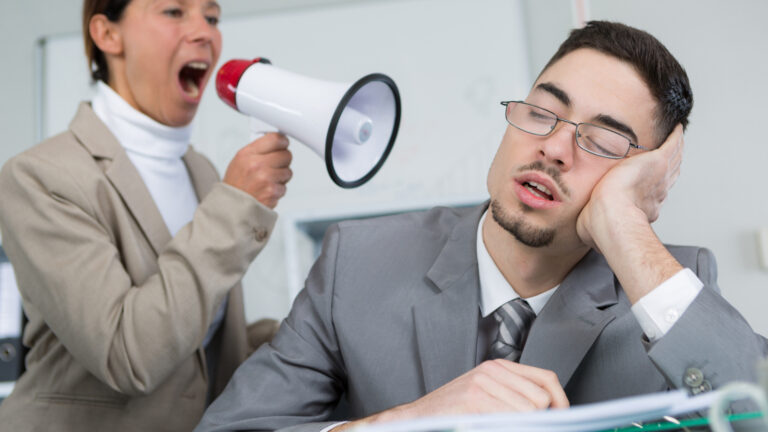 Woman in beige suit shouting through megaphone at sleeping male colleague in gray suit who rests his head on his hand, looking bored.