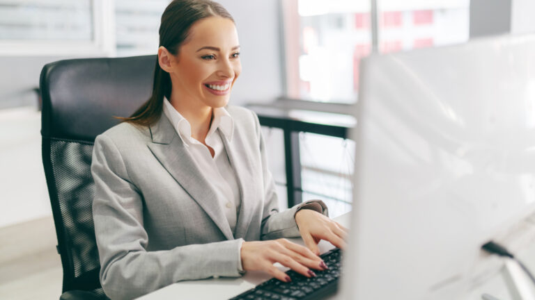 Smiling businesswoman in light gray suit working at computer in bright modern office, sitting in ergonomic chair.
