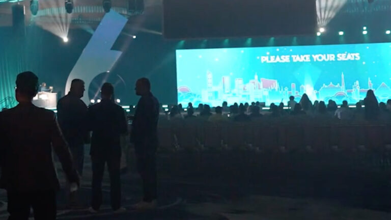 Darkened conference venue with audience silhouettes and large screen displaying "PLEASE TAKE YOUR SEATS" with city skyline.