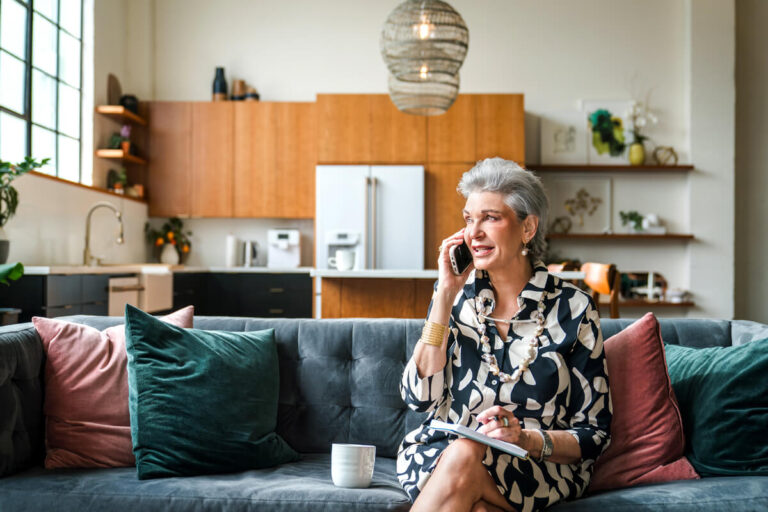 Woman with silver hair in patterned blouse sits on gray sofa with colorful pillows, talking on phone while holding tablet. Modern kitchen visible in background.