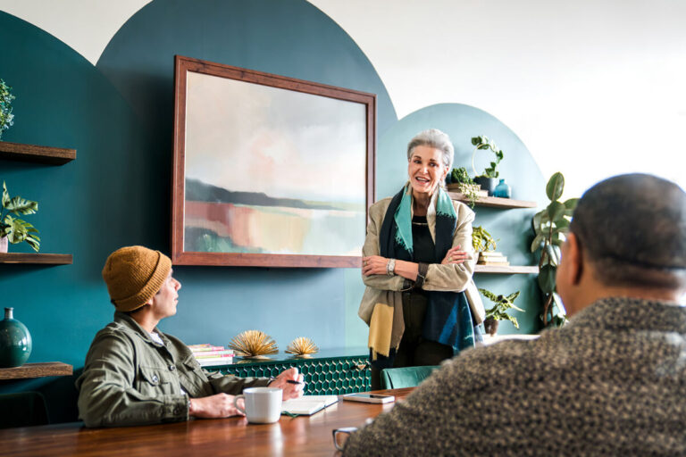 Three people conversing: silver-haired woman in beige blazer standing, person in yellow beanie seated with mug, another seated with back to camera. Teal walls with shelves.