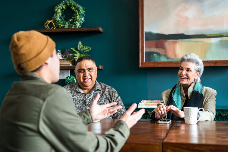 Three people at wooden table: person in yellow beanie gesturing, person in gray shirt listening, silver-haired woman in beige blazer smiling. Teal wall with shelves behind.