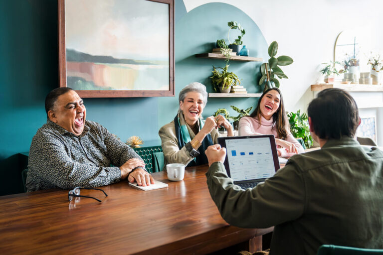 Four people at wooden table: person in gray shirt laughing, two women (silver-haired in blazer and dark-haired in pink) laughing, person showing laptop dashboard. Teal walls with shelves.