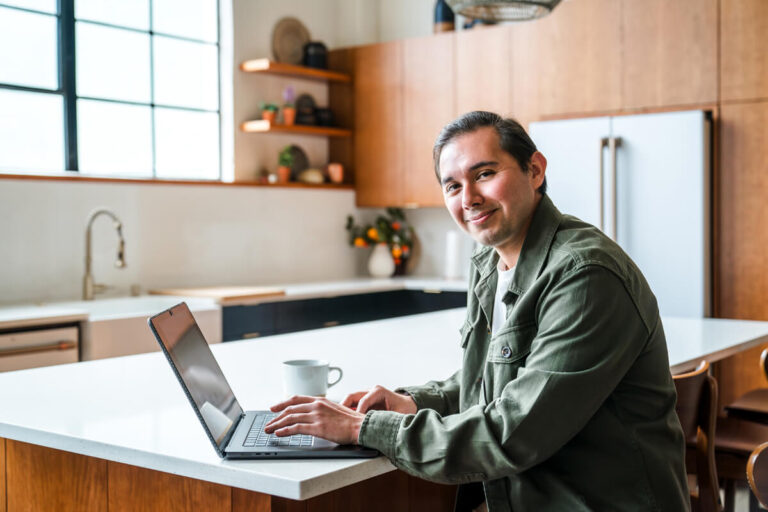 Smiling man in green jacket working on laptop at white kitchen counter with coffee mug, modern wooden cabinets in background.