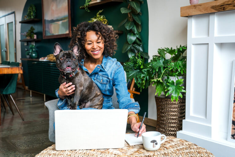 Smiling woman in denim shirt holding French bulldog while writing notes beside open laptop, surrounded by plants and teal accent wall.
