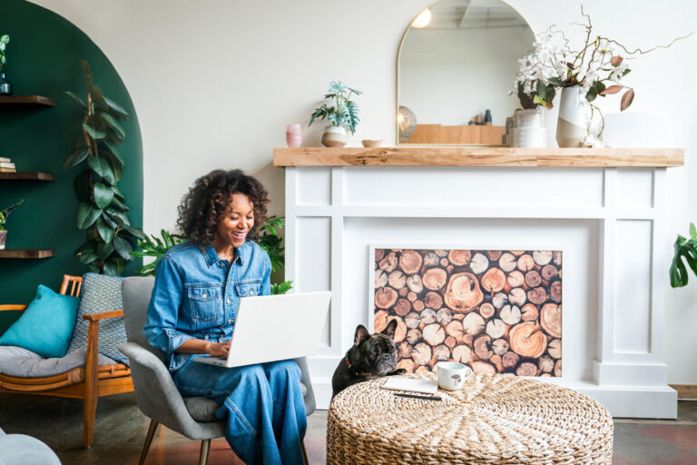 Woman in denim outfit using laptop in stylish living room with teal accent wall, French bulldog resting on wicker ottoman by decorative fireplace.