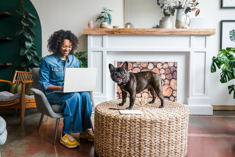 Woman in denim working on laptop while French bulldog stands on wicker ottoman, in living room with teal accent wall and decorative fireplace.