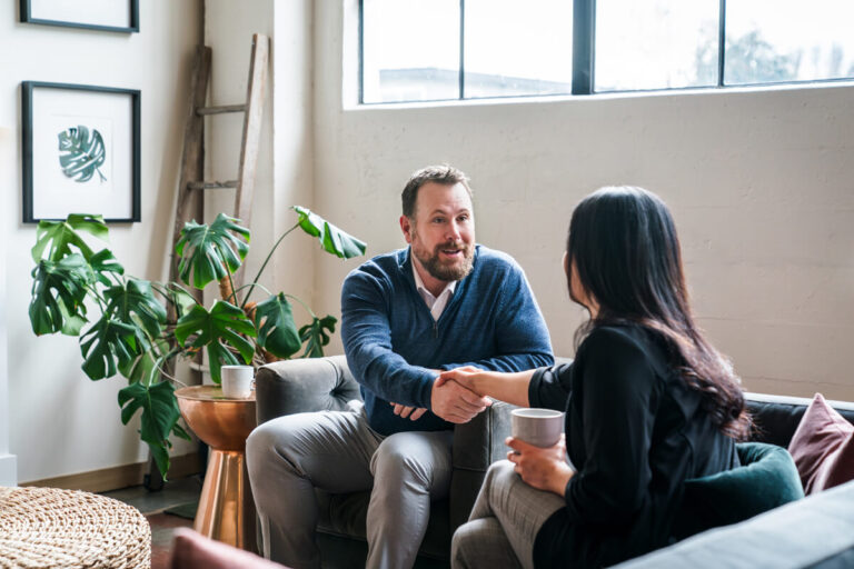 Two professionals shaking hands in a modern lounge. Bearded man in blue sweater meets woman in black top with coffee mug. Bright space with plants and contemporary decor.