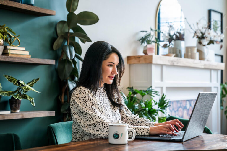 AWoman with dark hair works on laptop at wooden table. Teal chair, white mug, plant-filled office with teal accent wall and decorative shelving.