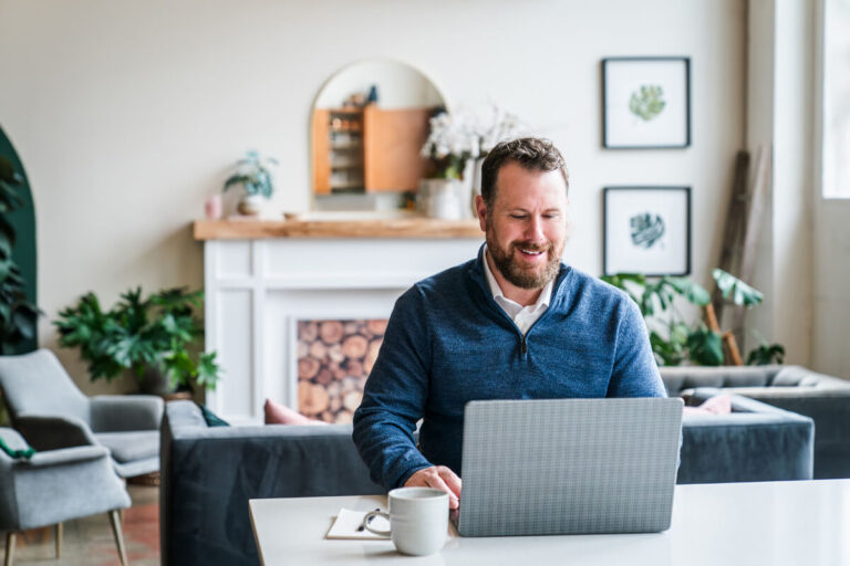 A bearded man in blue sweater smiles while working on laptop at white table with coffee mug. Stylish background with fireplace, wooden mantle, plants, and gray furniture.