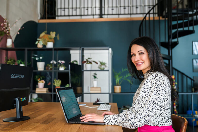 Woman with dark hair smiles at camera, working at desk with laptop and monitor. White/black blouse, pink pants. Teal wall, spiral staircase, plant shelves.