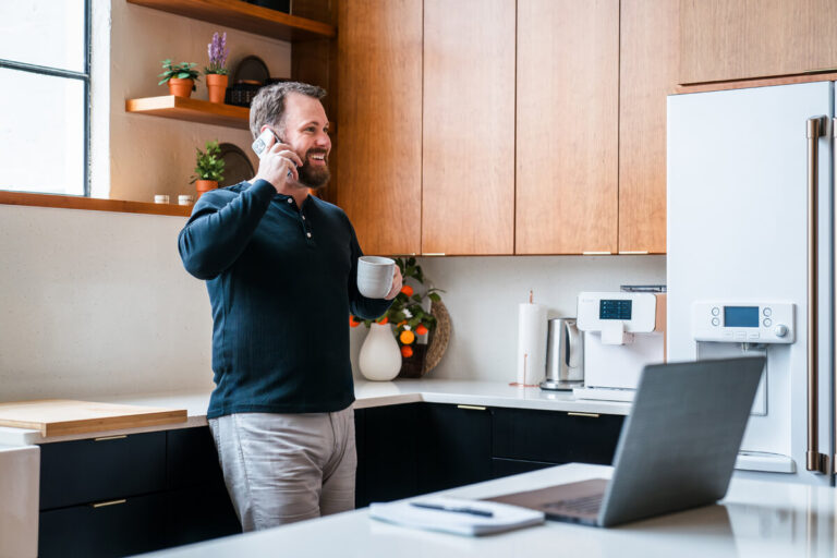 A bearded man in navy henley smiles while talking on phone and holding a mug in modern kitchen with wooden/black cabinets, white countertops, and laptop on island.