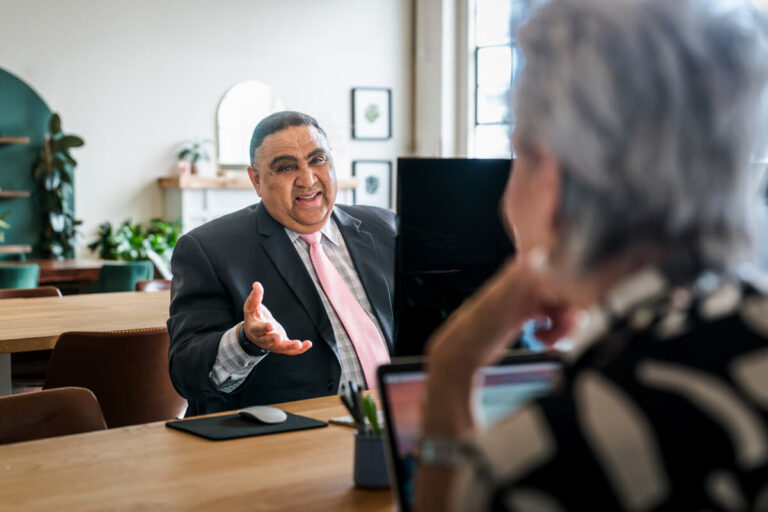 A person with gray hair in patterned outfit meets with a man in dark suit and pink tie who gestures while speaking across wooden table in bright modern office with plants and artwork.