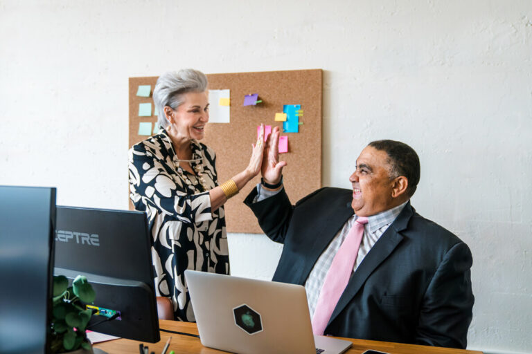 Two colleagues high-fiving in office: silver-haired woman in patterned blouse with seated man in dark suit and pink tie. Cork board and laptop visible.